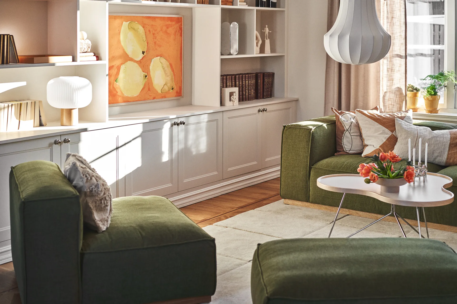 Ηλιόλουστο σαλόνι με πράσινους καναπέδες, ένα λευκό τραπεζάκι με πορτοκαλί τουλίπες και κεριά, και λευκά ράφια με τέχνη.