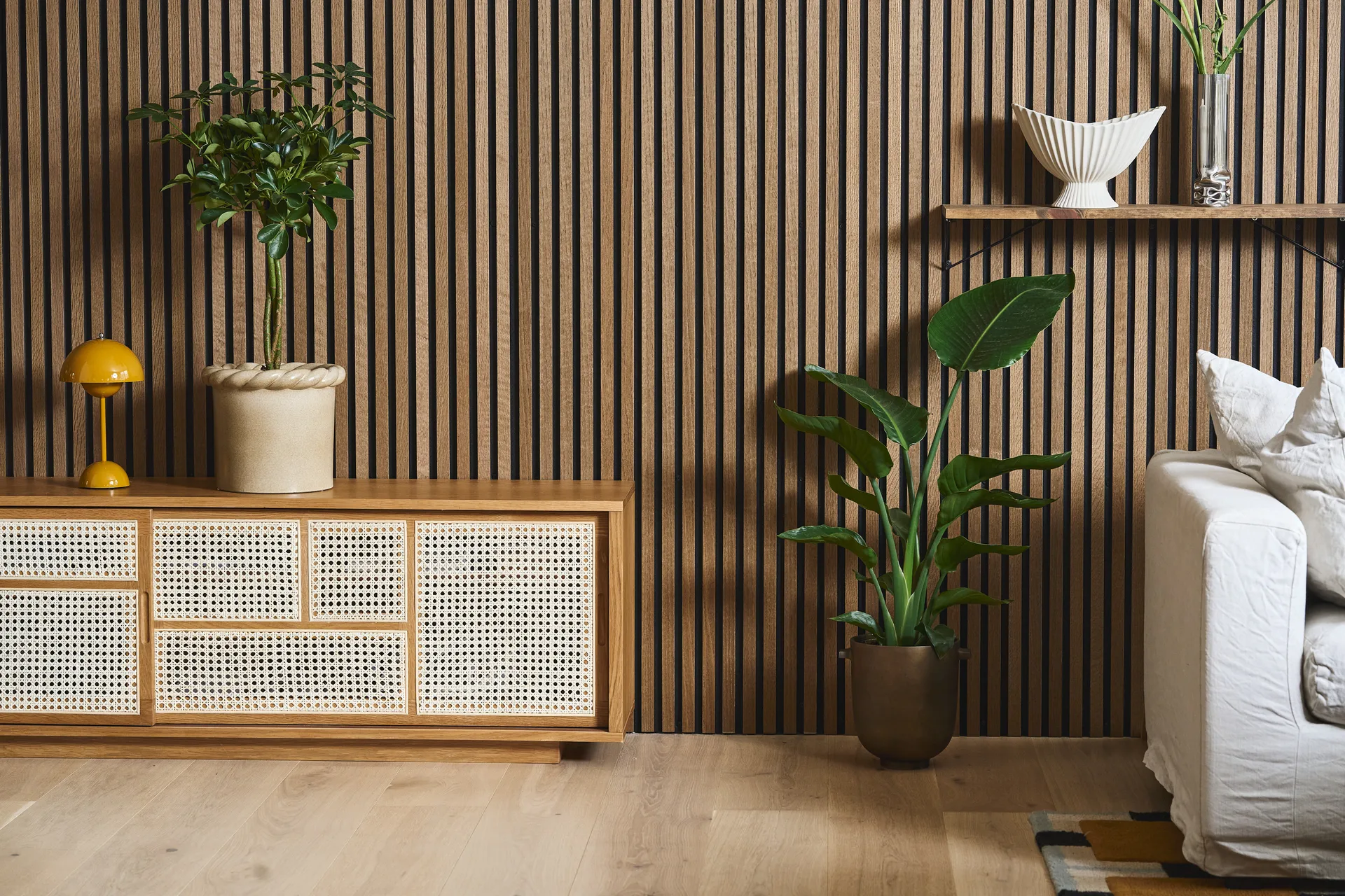 Μοντέρνο σαλόνι με τοίχο από ξύλινες σανίδες, ντουλάπι από ρατάν, κίτρινο φωτιστικό, φυτά και λευκό καναπέ.