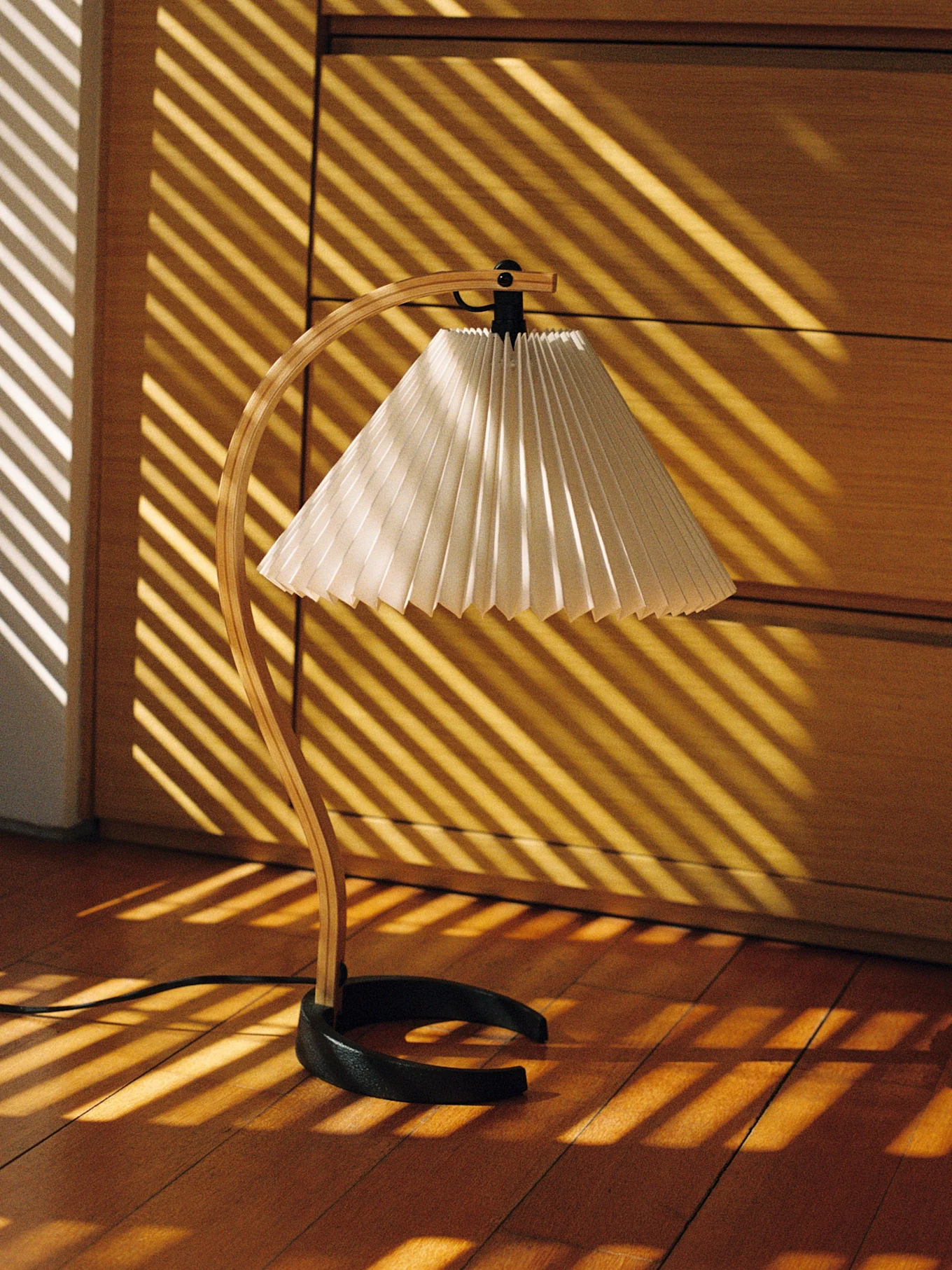 Επιτραπέζιο φωτιστικό με ξύλινη βάση και πλισέ αμπαζούρ, με ρίγες φωτός στο ξύλινο πάτωμα.