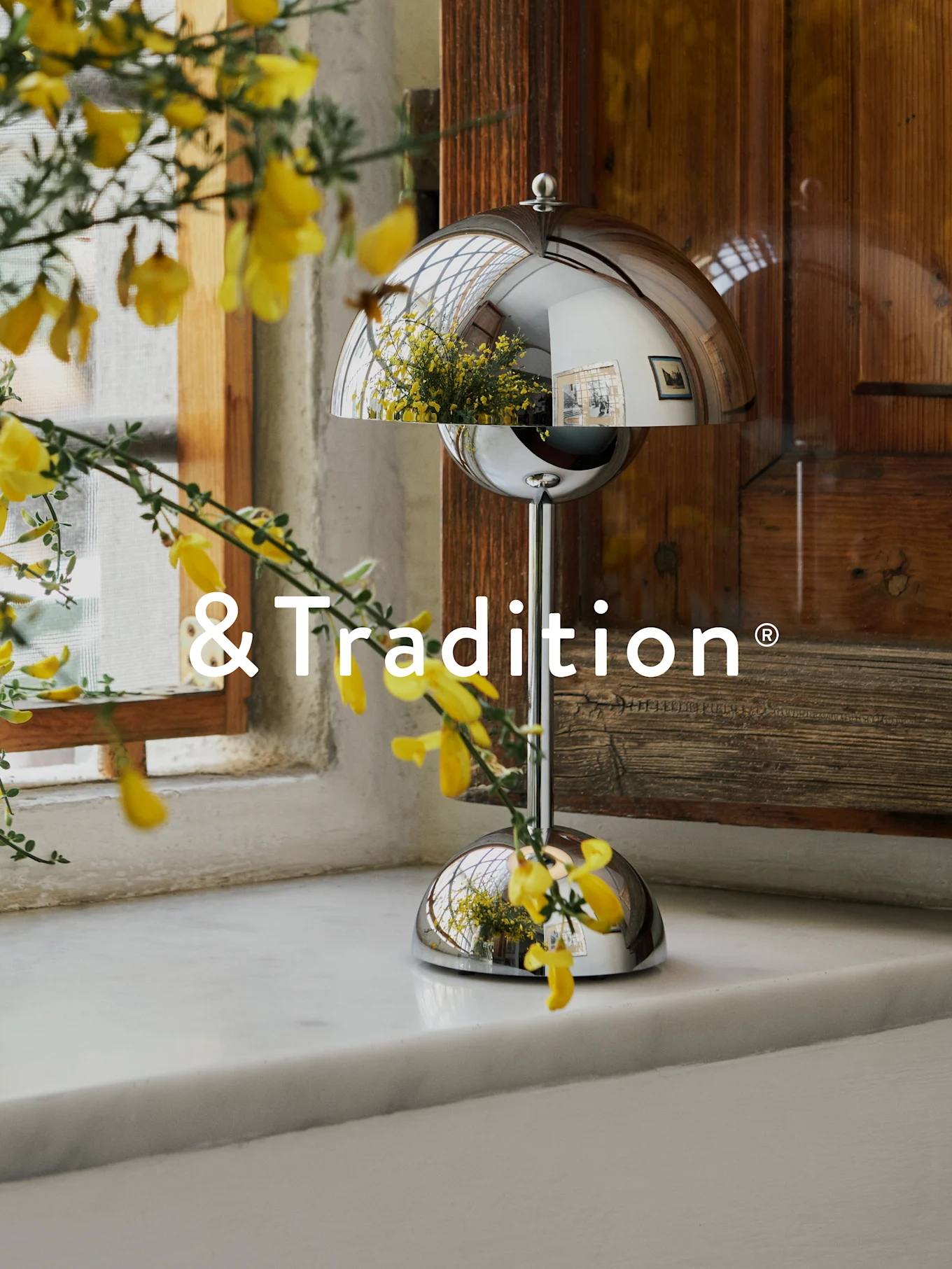 Ένα ασημένιο, θολωτό επιτραπέζιο φωτιστικό σε λευκό περβάζι με κίτρινα λουλούδια μπροστά. Το φωτιστικό αντανακλά το παράθυρο και τα λουλούδια. Λογότυπο '&Tradition'.