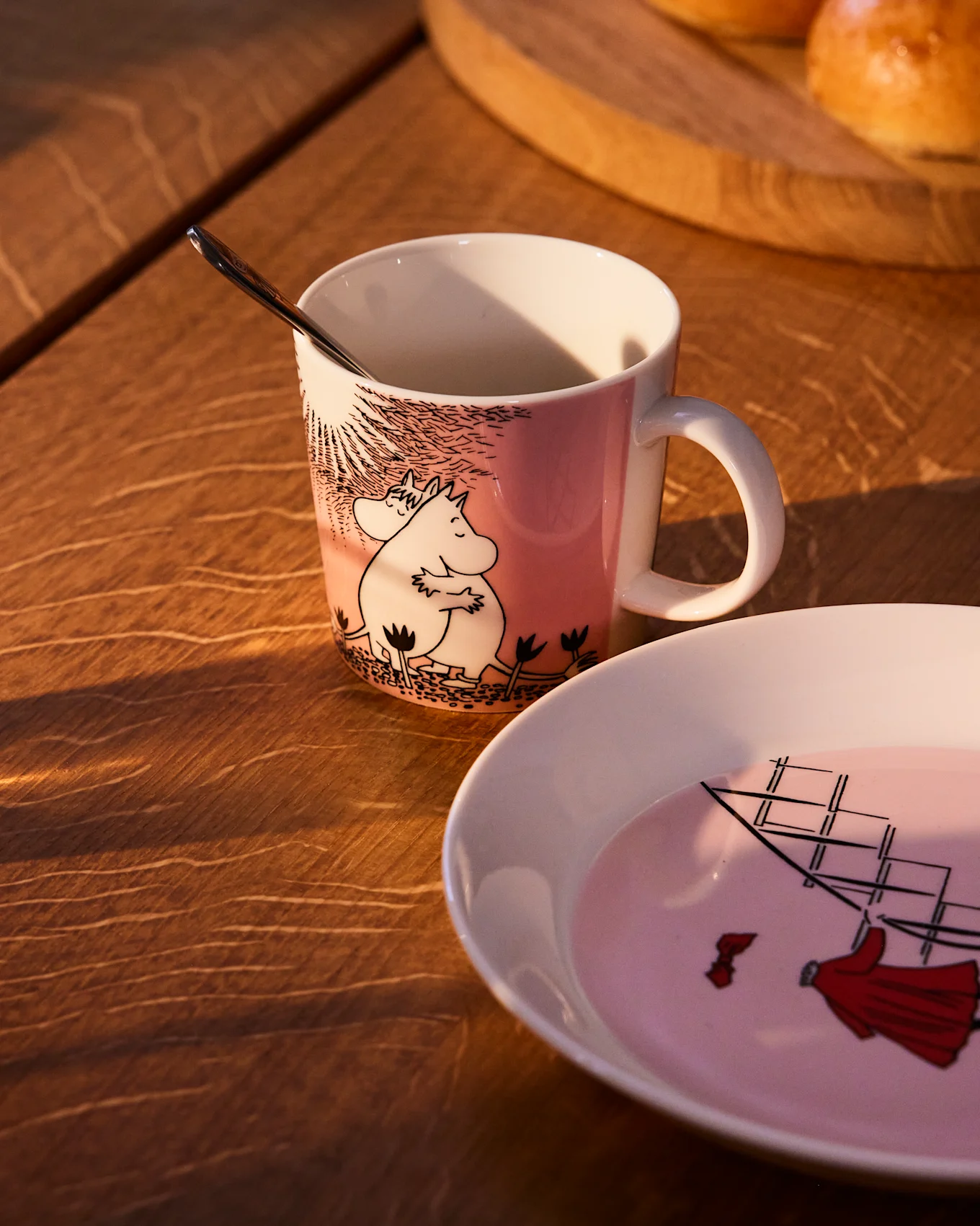 Ροζ κούπα Moomin με τον Moomintroll και τη Snorkmaiden, δίπλα σε ένα ασορτί πιάτο με ιστιοφόρο, πάνω σε ένα ξύλινο τραπέζι.
