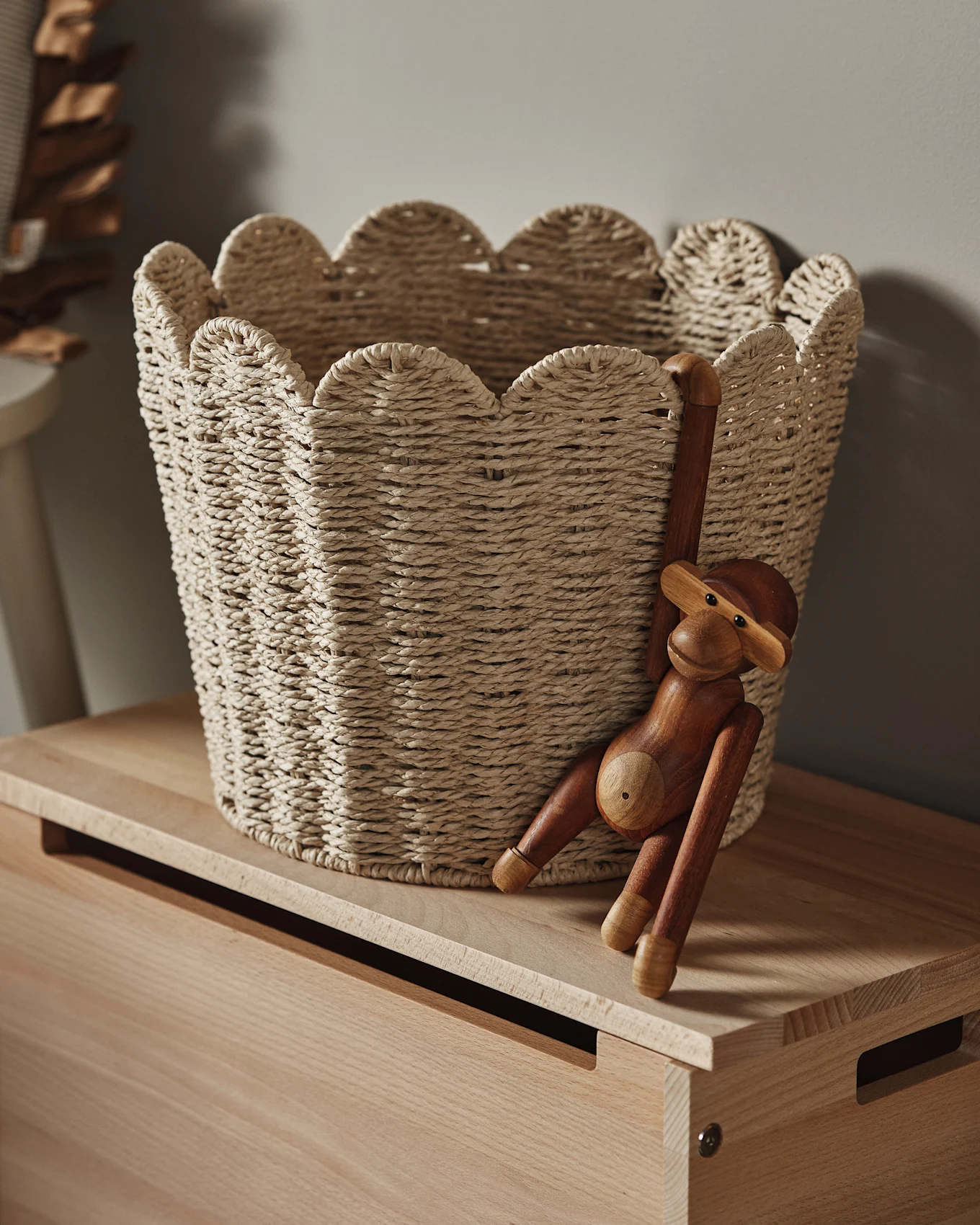 Ένα ανοιχτόχρωμο πλεκτό καλάθι με κυματιστό χείλος κάθεται σε μια ξύλινη επιφάνεια, με ένα ξύλινο ειδώλιο μαϊμού να κρέμεται από την πλευρά του.