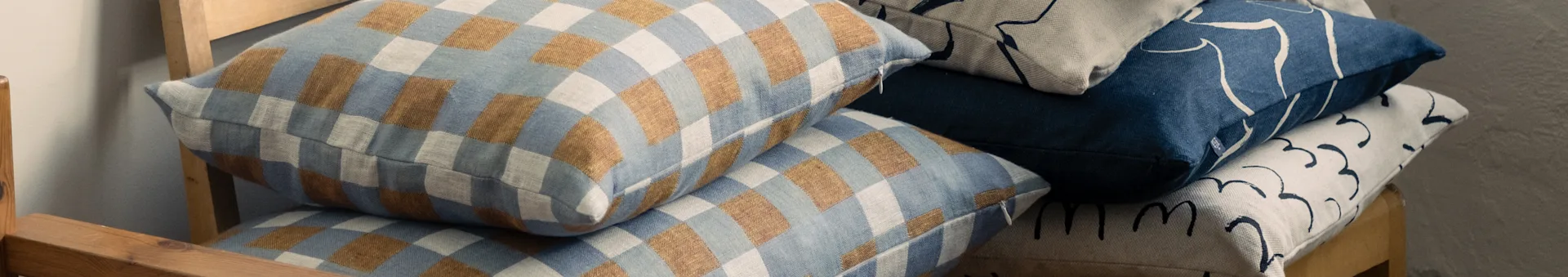 Στοιβασμένα μαξιλάρια σε ξύλινη καρέκλα, με καρό μπλε και καφέ, και αφηρημένα σχέδια σε μπλε και μπεζ αποχρώσεις.