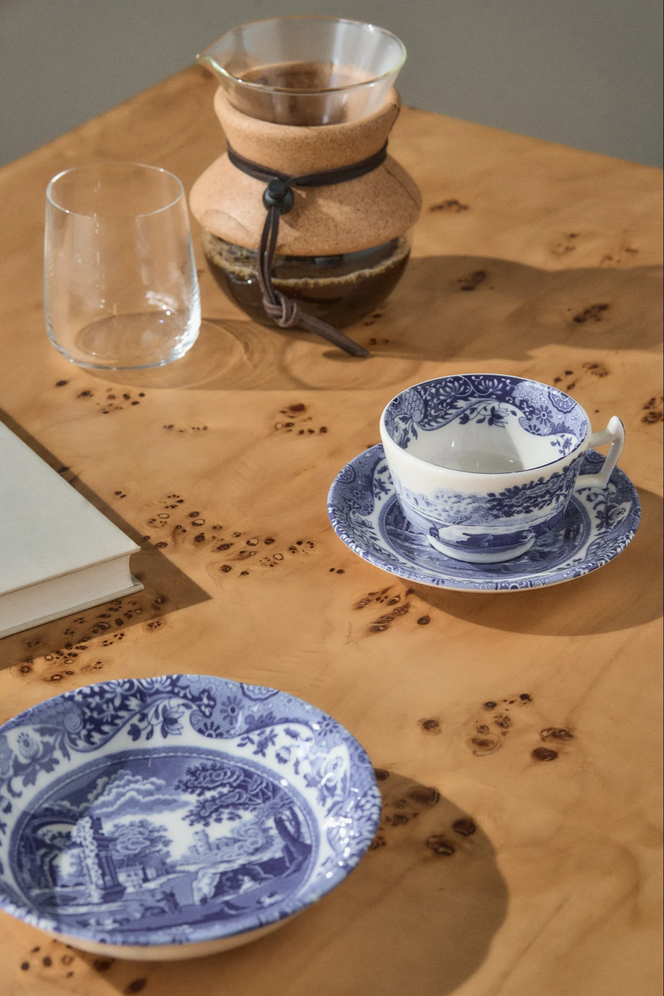 Μια καφετιέρα pour-over, ένα άδειο ποτήρι, ένα μπλε και λευκό φλιτζάνι τσαγιού σε πιατάκι, και ένα ασορτί πιάτο σε ένα ανοιχτόχρωμο ξύλινο τραπέζι.