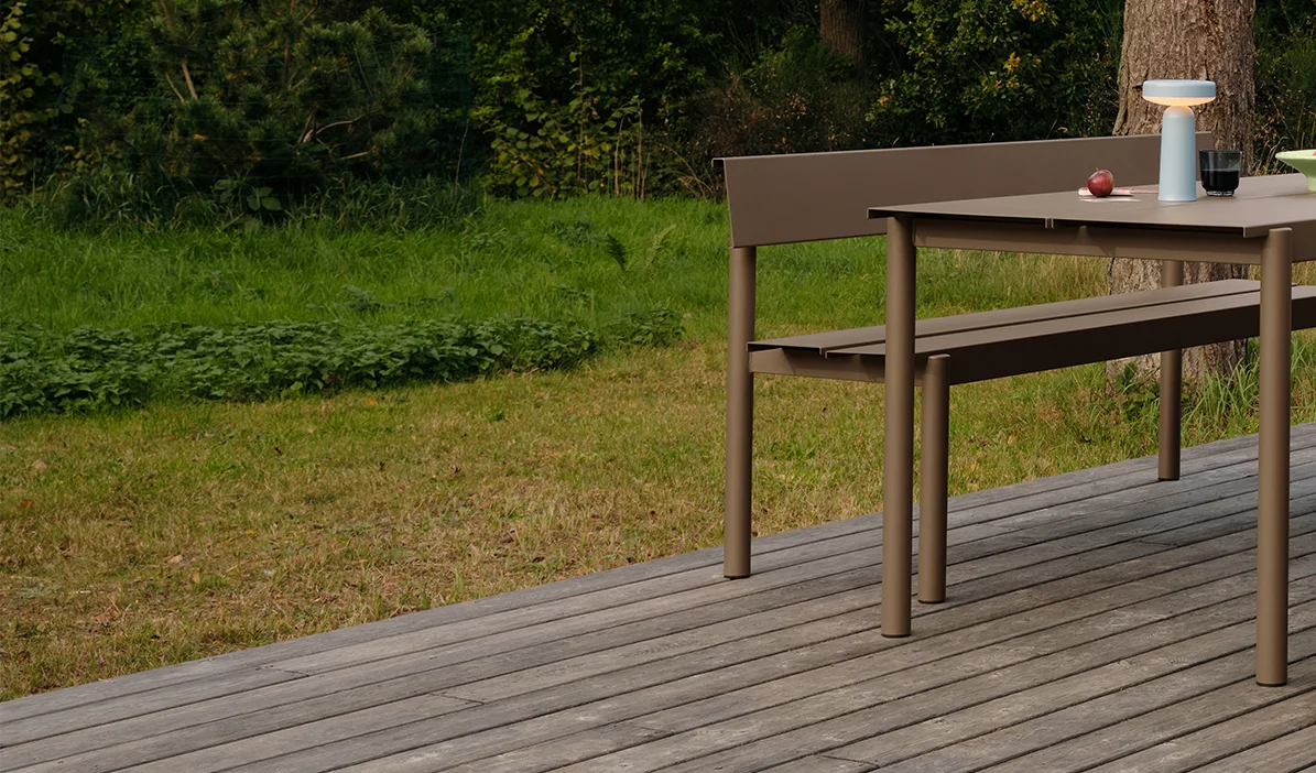 Μοντέρνο τραπέζι και πάγκος εξωτερικού χώρου σε χρώμα μόκα σε ξύλινο κατάστρωμα, με φορητό φωτιστικό και μήλα στο τραπέζι.