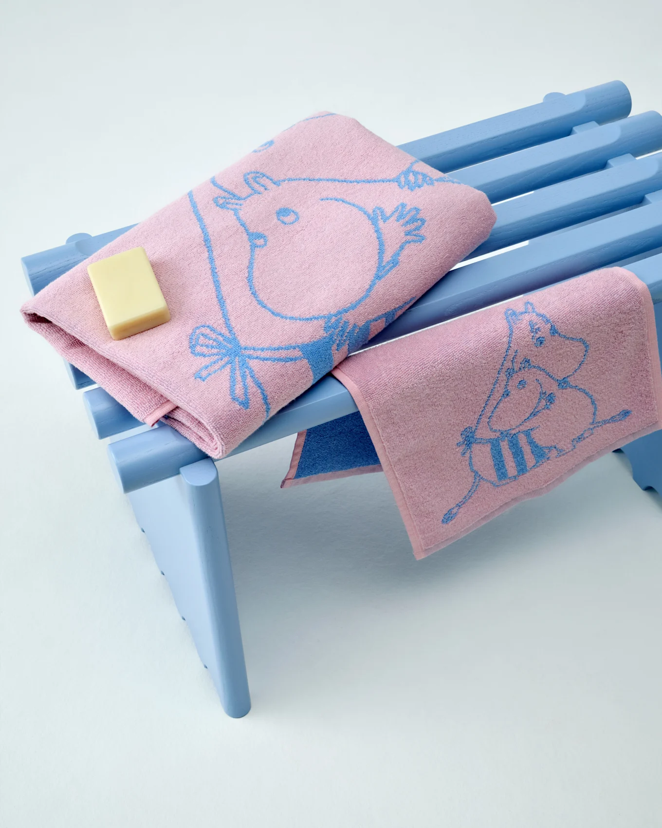 Δύο ροζ πετσέτες Moomin με μπλε σχέδια, η μία διπλωμένη με μια μπάρα σαπουνιού, σε ένα γαλάζιο παγκάκι.