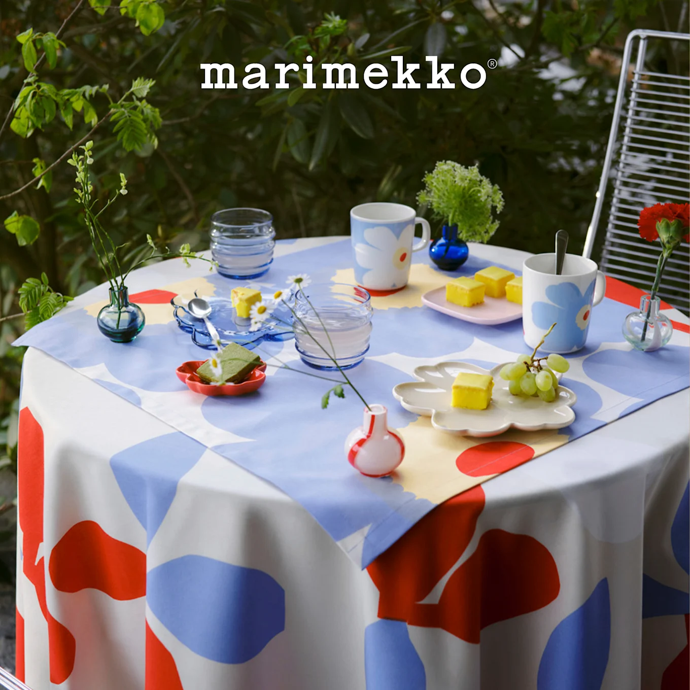 Εξωτερικό τραπέζι με τραπεζομάντιλο Marimekko με κόκκινο, μπλε, λευκό σχέδιο, στρωμένο με κούπες με λουλούδια, επιδόρπια και μικρά φυτά. Λογότυπο Marimekko.