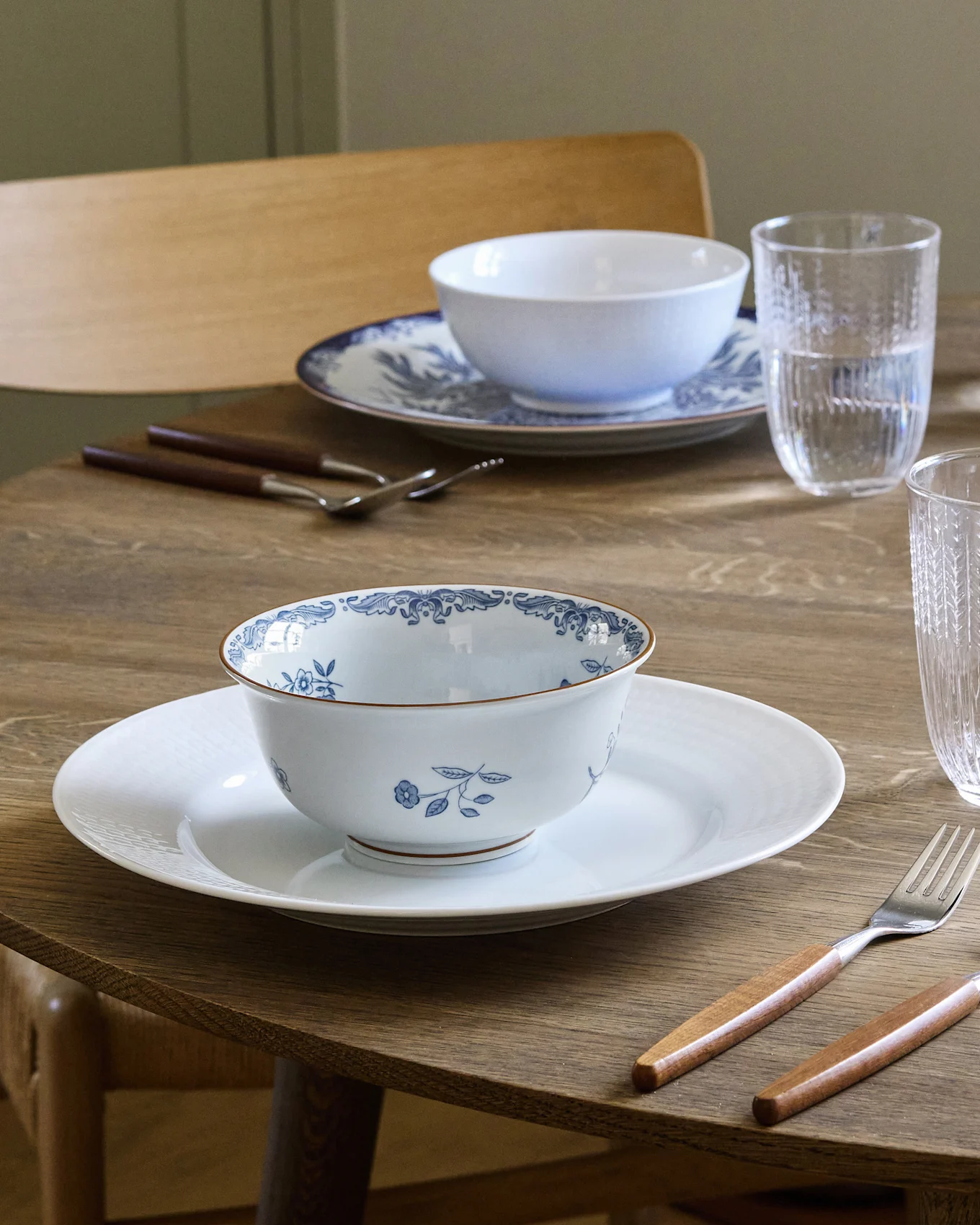 Τραπέζι φαγητού στρωμένο για δύο με λευκά και μπλε μπολ με σχέδια σε λευκά πιάτα, μαχαιροπίρουνα με ξύλινες λαβές και ποτήρια νερού.