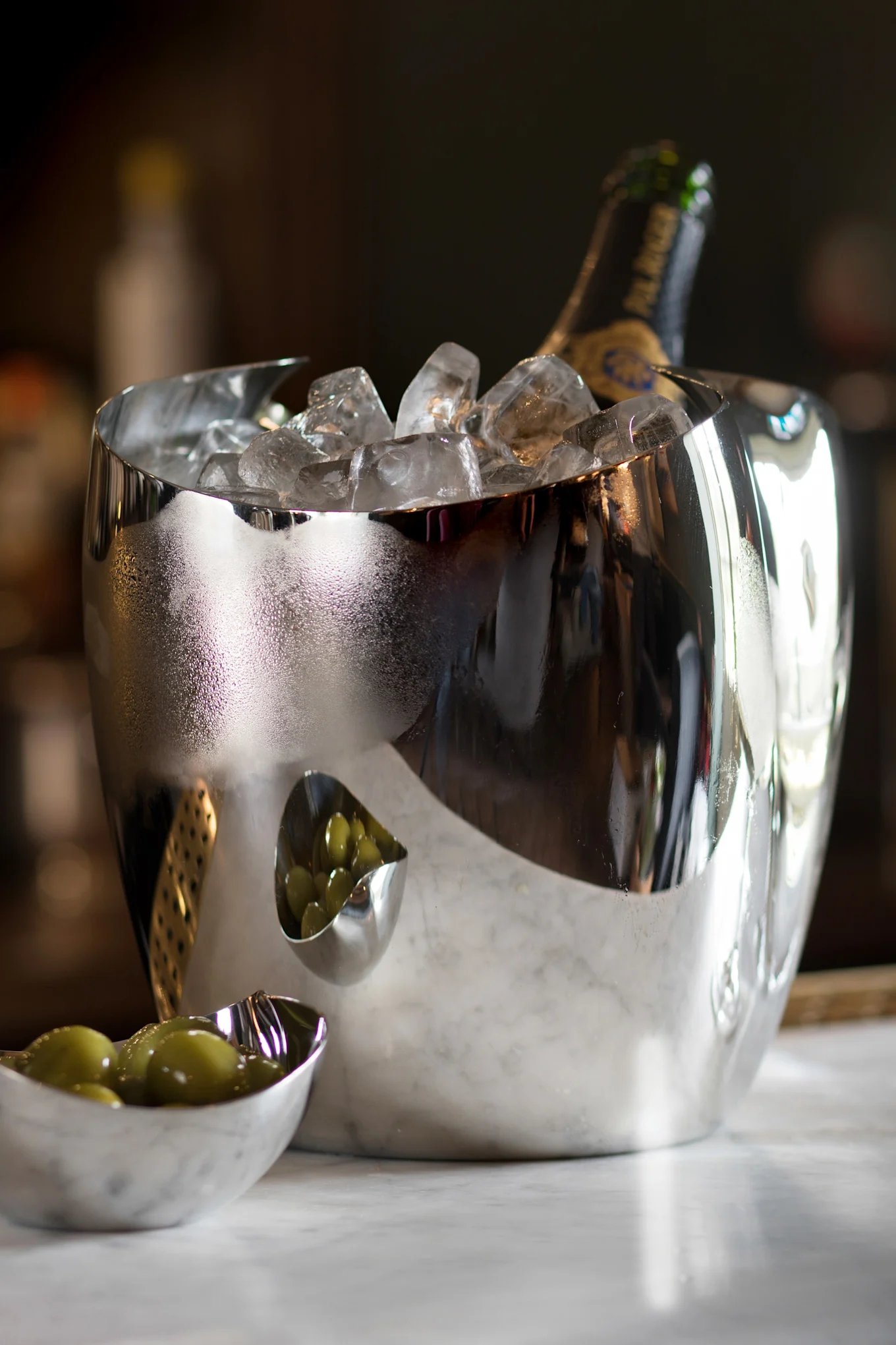 Ένας ανακλαστικός ασημένιος κάδος πάγου με μπουκάλι σαμπάνιας και πάγο, δίπλα σε δύο μικρά μπολ με πράσινες ελιές σε μάρμαρο.