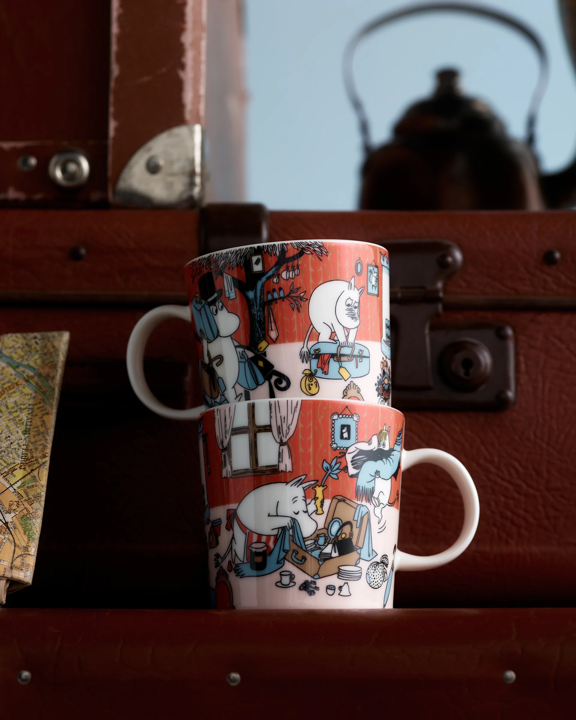 Δύο κούπες Moomin με πορτοκαλοκόκκινες εικονογραφήσεις χαρακτήρων, στοιβαγμένες μπροστά από καφέ βαλίτσες και έναν θολό χάρτη.