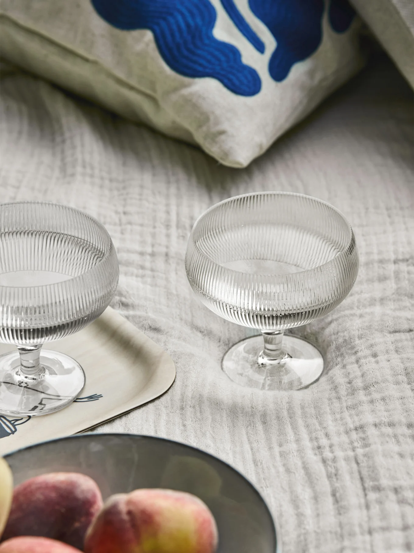 Δύο ραβδωτά γυάλινα μπολ επιδόρπιου, ένα μαξιλάρι με μπλε αφηρημένη τέχνη, ένας ξύλινος δίσκος και ροδάκινα σε ανοιχτόχρωμο ύφασμα.