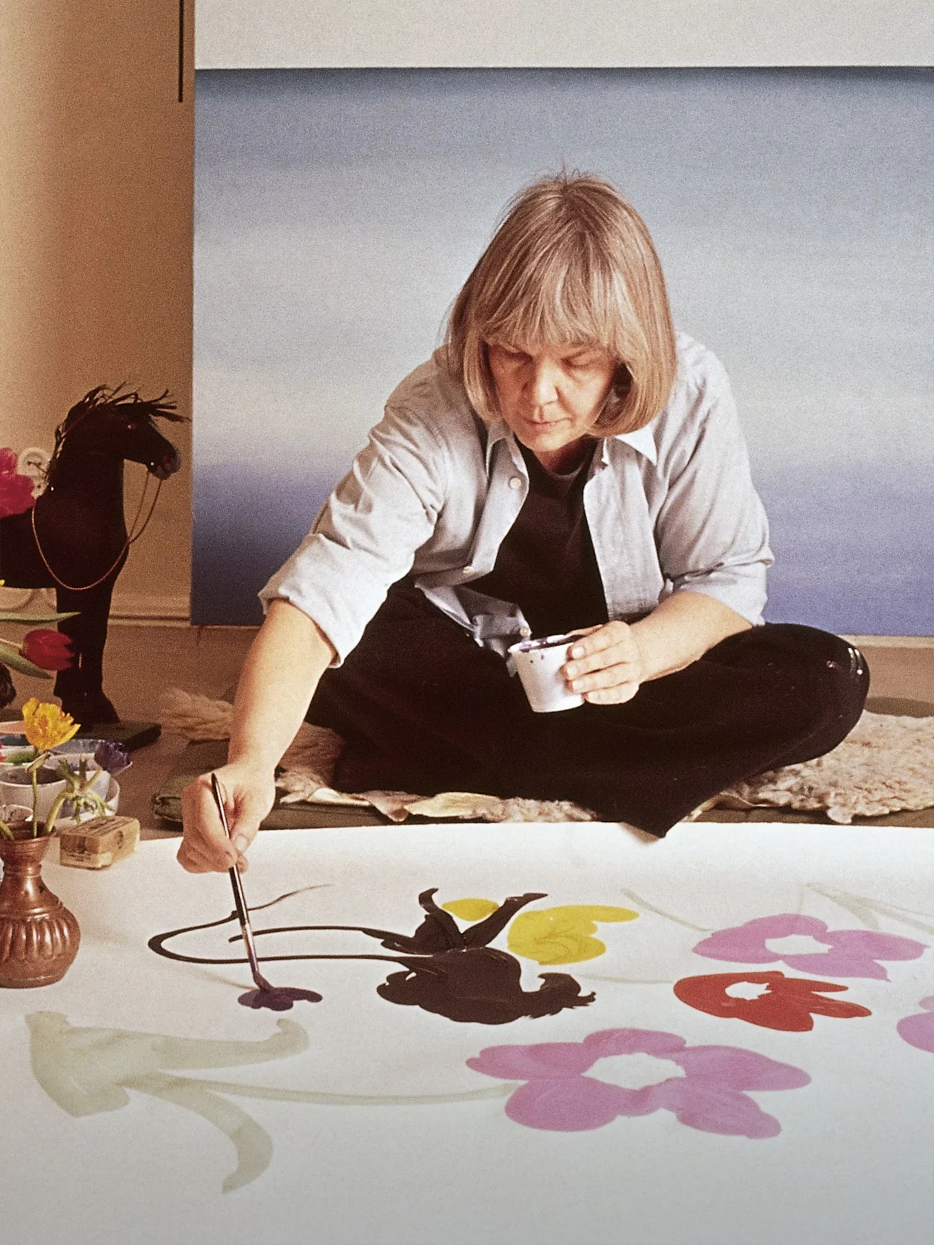Η καλλιτέχνις Marimekko Maija Isola κάθεται στο πάτωμα και ζωγραφίζει μεγάλα, πολύχρωμα λουλούδια σε ένα λευκό ύφασμα.
